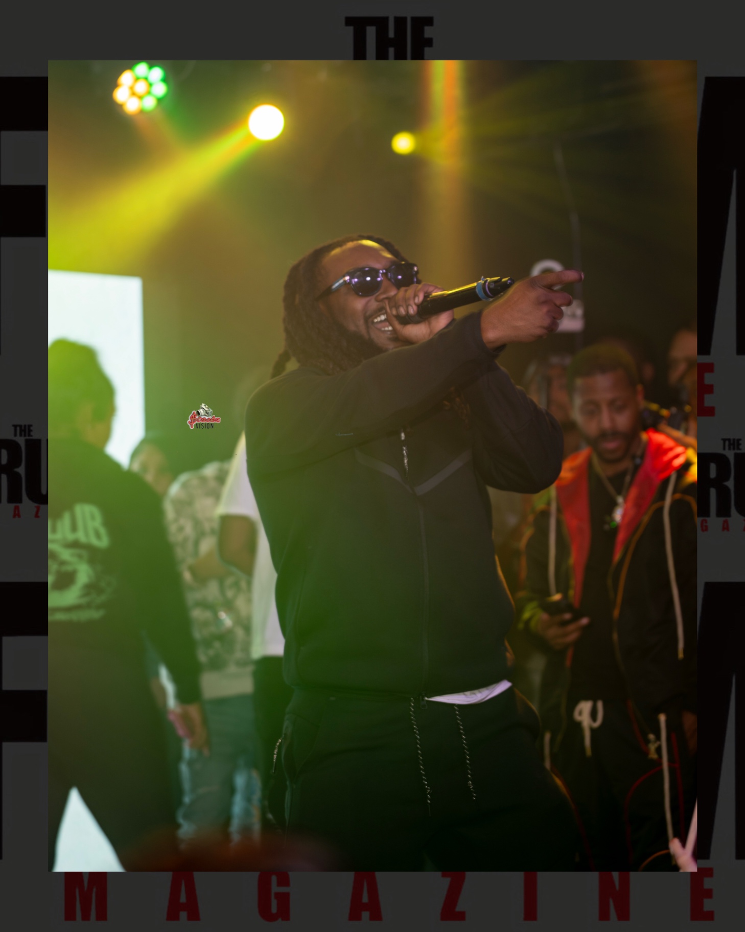 A performer pointing toward the crowd while rapping into a microphone under green and yellow lights
